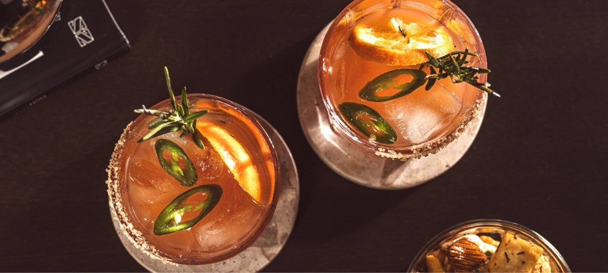 Drei elegante Cocktailgläser mit bernsteinfarbenem Getränk, Jalapeño-Scheiben und Rosmaringarnituren auf dunklem Hintergrund