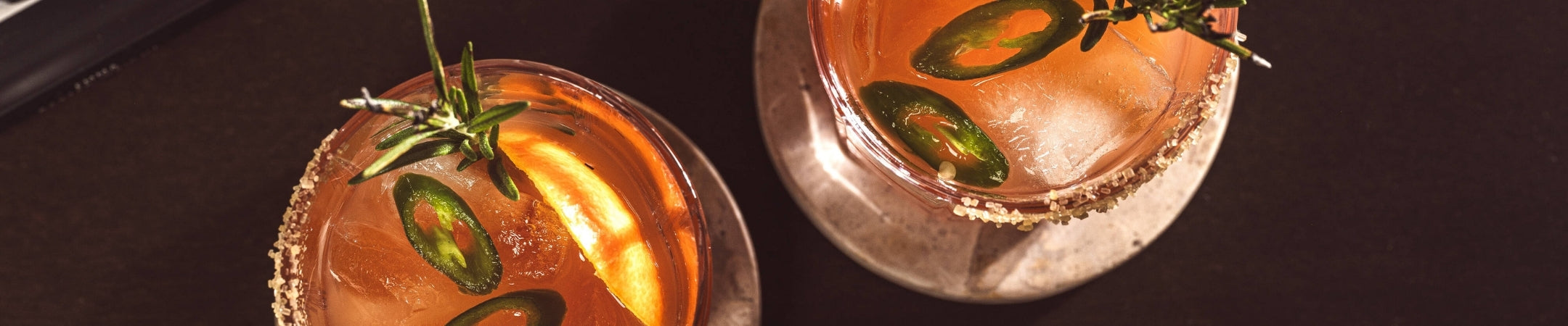 Zwei Cocktails mit Jalapeno-Scheiben und Rosmarin-Garnierung auf dunklem Hintergrund, warm-orange beleuchtet
