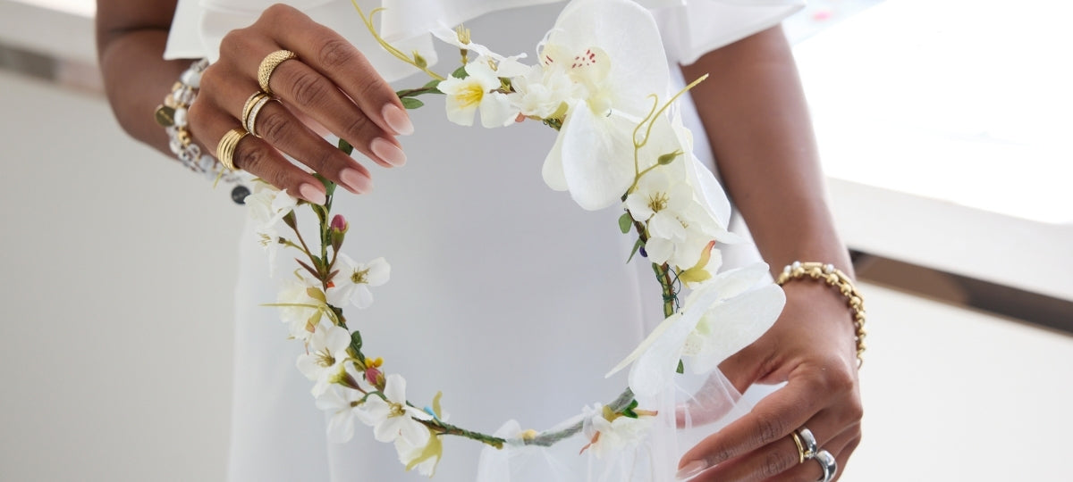 Eine Frau in einem weißen Kleid hält einen filigranen Blumenkranz mit weißen Blüten in den Händen und trägt mehrere goldene Ringe sowie ein Armband, die den eleganten, festlichen Look unterstreichen.