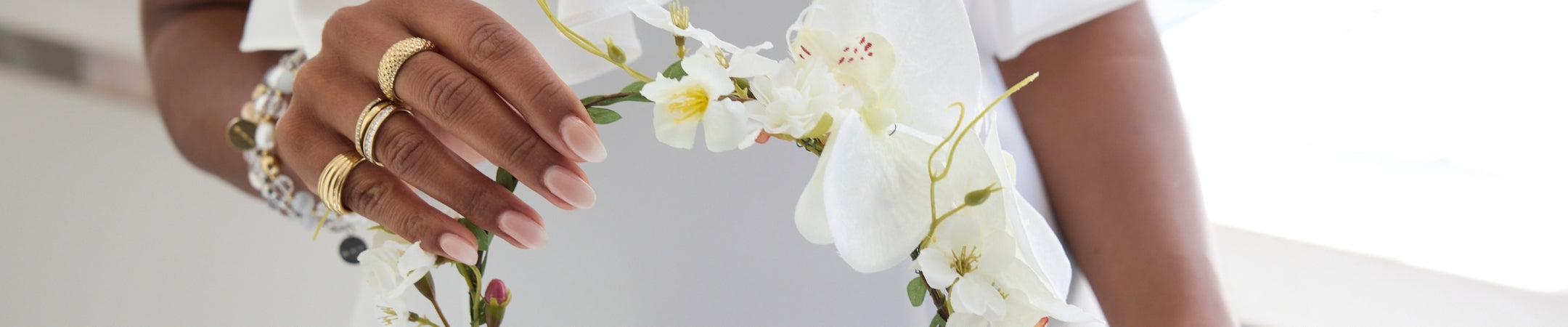 Detailaufnahme einer Frau in weißer Kleidung, die einen zarten Blumenkranz aus weißen Blüten hält, während goldene Ringe und ein Armband ihre Hände schmücken und eine stilvolle, festliche Atmosphäre schaffen.