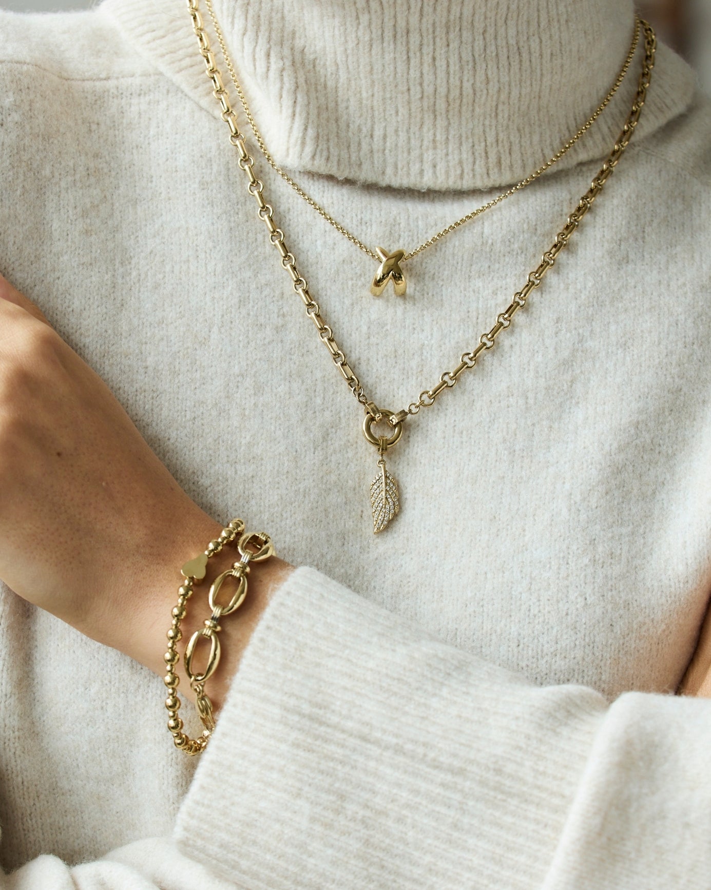 Goldene Kettenhalsketten mit Anhängern und passende Armbänder an Frauenhand, cremefarbener Pullover