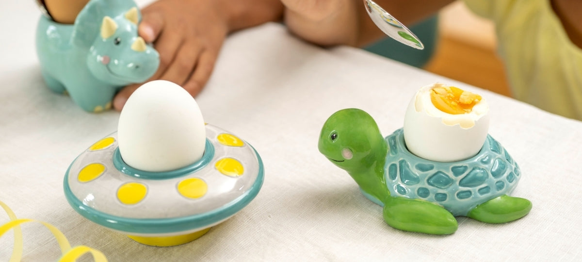 Bunte Kinder-Eierbecher: UFO mit gelbem Muster und grüne Schildkröte halten weiße Eier, daneben Spielzeug-Dinosaurier