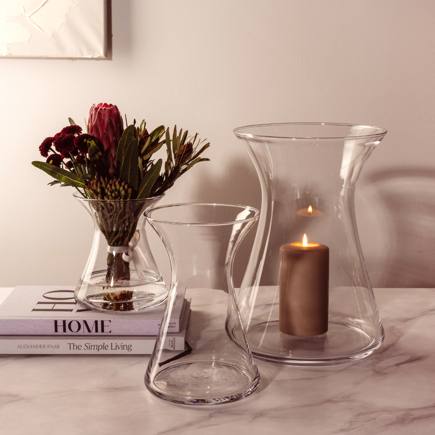 Elegante Glasvasen mit roten Blumen, brennenden Kerzen und Designbüchern auf Marmortisch in warmer Atmosphäre