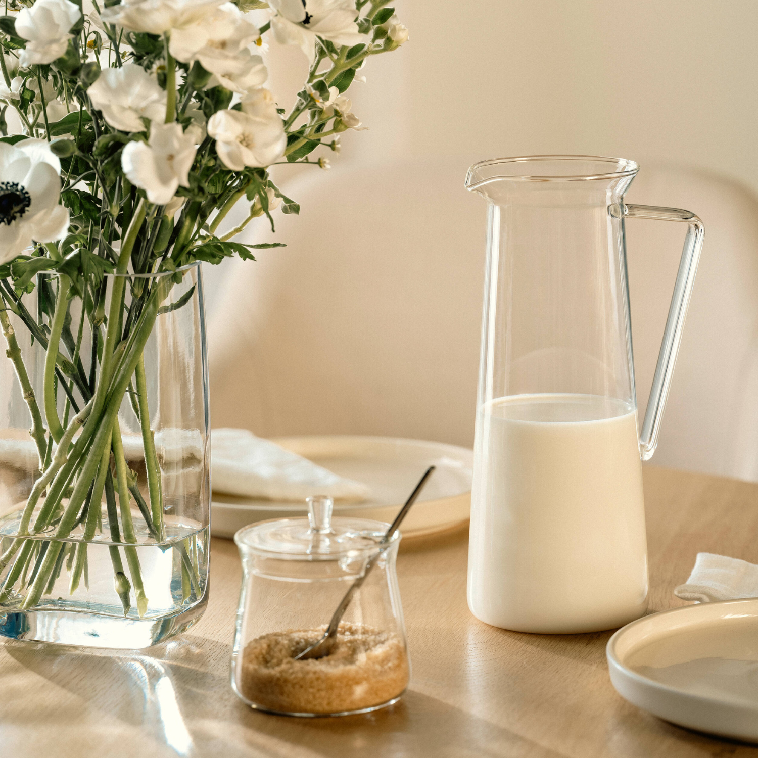Elegante Tischszene mit Glaskrug Milch, Blumenvase weiße Nelken, Zuckerdose und Tellern in warmem Beige