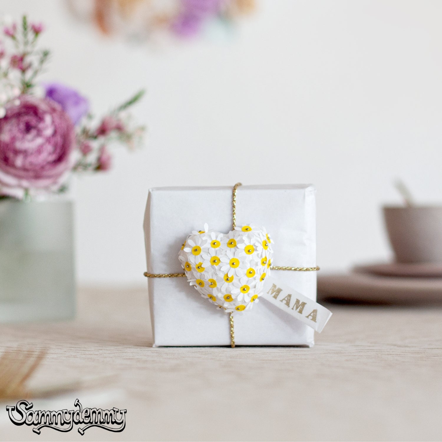 MAMA, weiß verpacktes Geschenk mit goldenem Bindfaden und herzförmigem Charm aus weißen Blüten mit gelben Zentren, florale Dekoration im Hintergrund