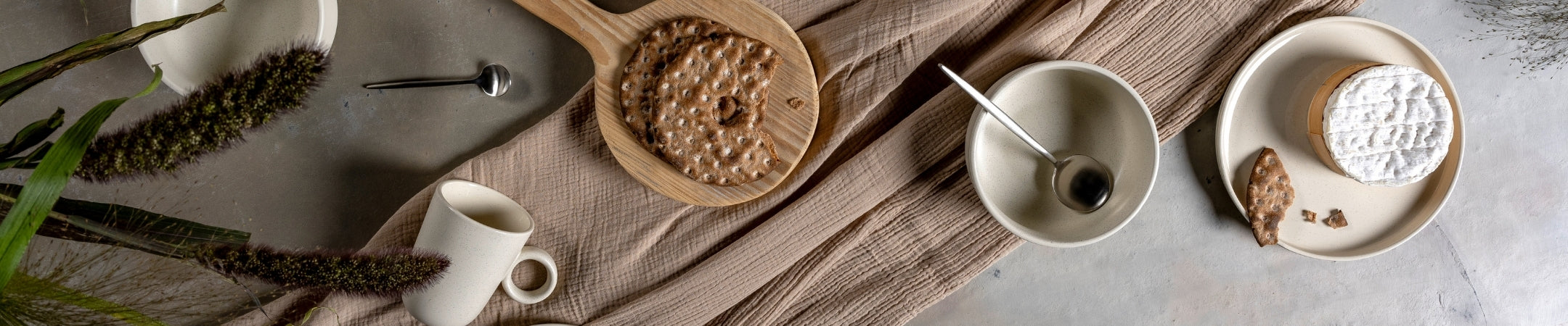 Flatlay mit Keramikschüsseln, Tellern, Besteck und Getreideähren auf grauem Leinen und heller Oberfläche