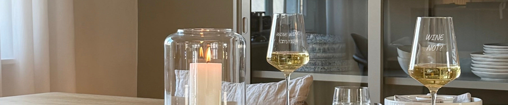 Elegante Weingläser mit Gravur auf hellem Marmor, gefüllt mit Wein, neben Kerze und Tellerstapel