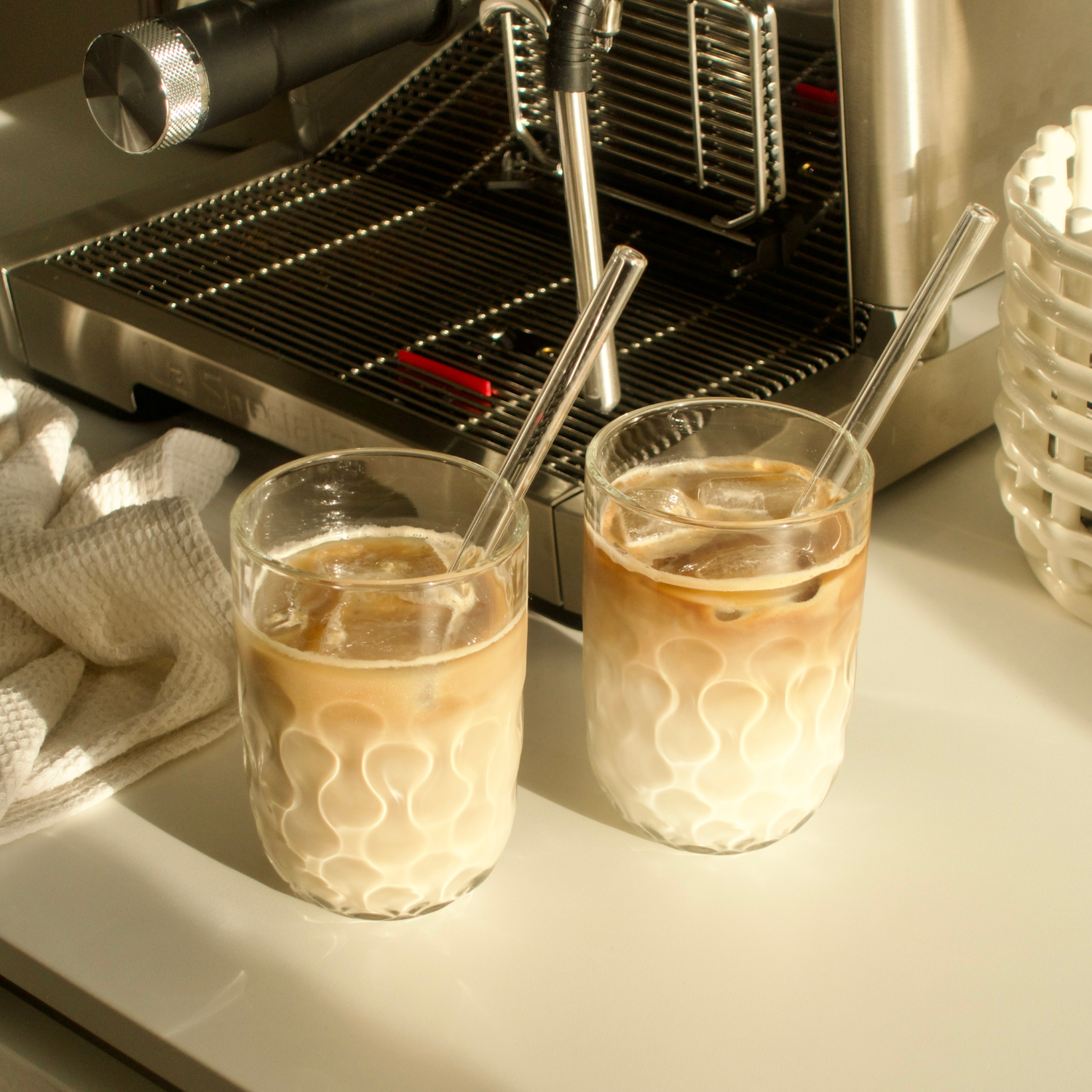 Zwei Gläser mit Eiskaffee und Strohhalmen vor einer Espressomaschine, warme Lichtbrechung