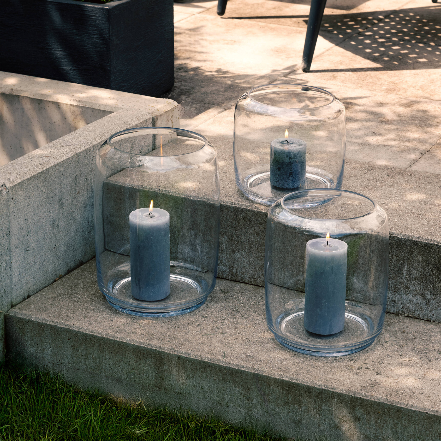 Vier blaue Kerzen in transparenten Glaszylinder-Behältern auf Betonpodest, sonnige Außenumgebung, moderne Deko-Arrangement