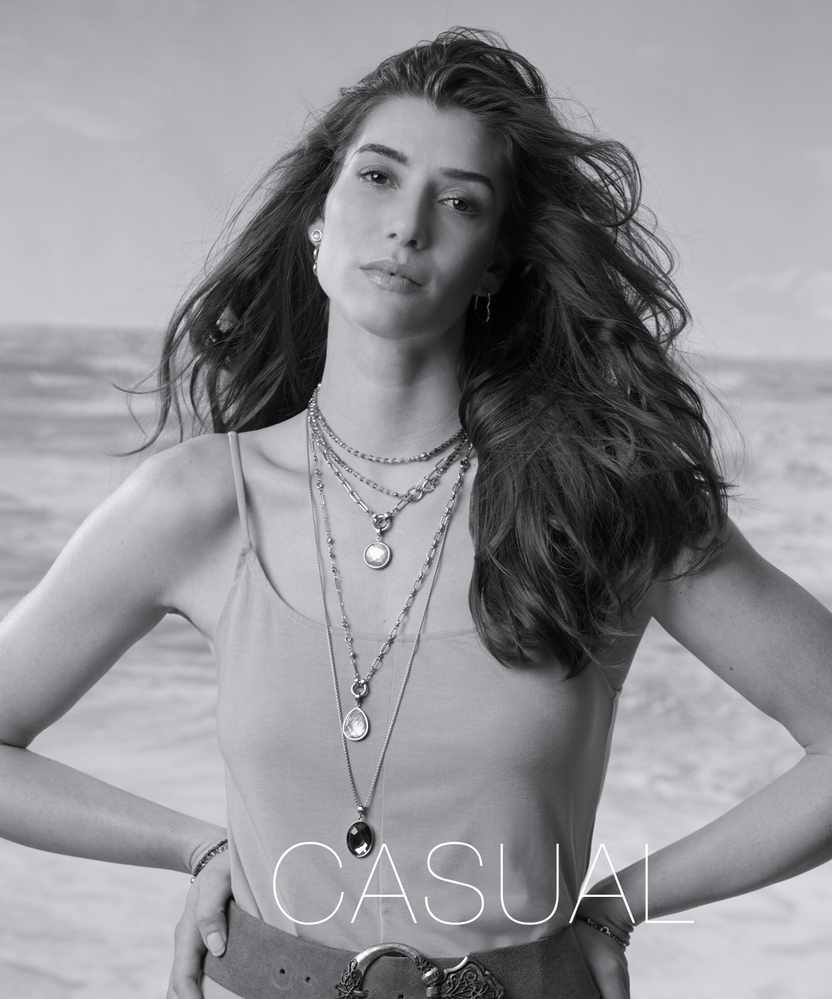 Eine Frau steht am Strand mit wehendem Haar und trägt mehrere filigrane Ketten im Layering-Stil. Sie blickt selbstbewusst in die Kamera und präsentiert modernen Schmuck in einem lässigen Look.