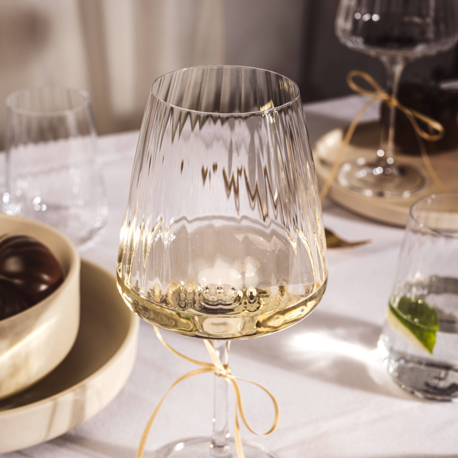 Elegantes Weinglas PAOLO mit gerilltem Design, gefüllt mit Weißwein, auf gedecktem Tisch mit goldener Schleife