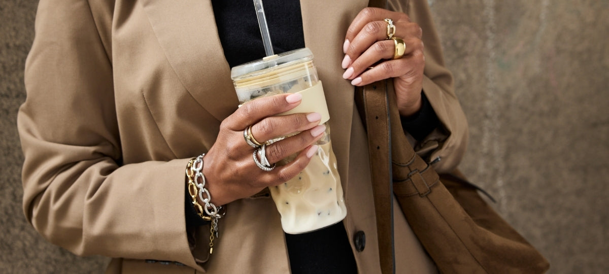 Frau trägt mehrere silberne und goldene Ringe an den Händen, hält Glaskanne mit Getränk, beige Jacke