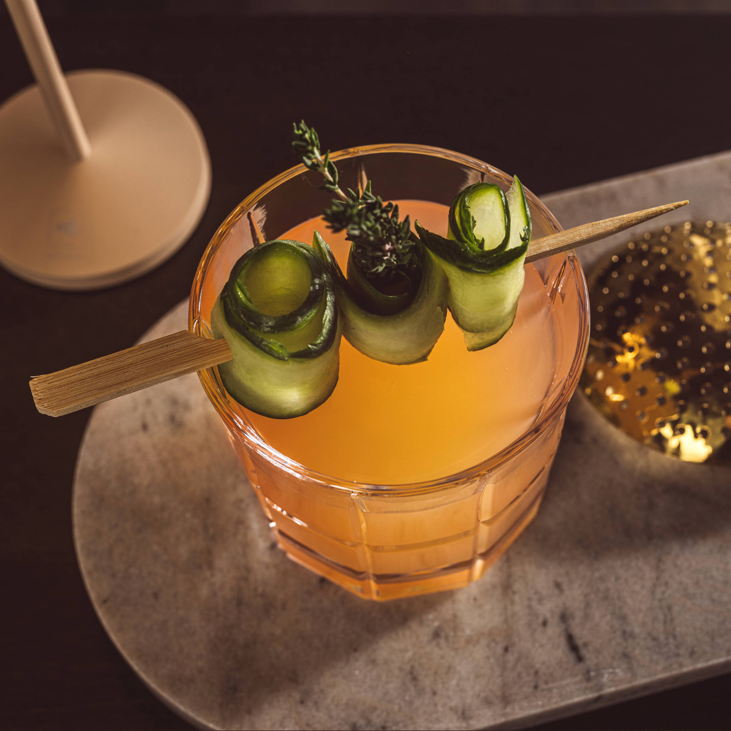 Elegantes Cocktailglas mit bernsteinfarbenem Drink, garniert mit Gurkenscheiben und Thymian auf Holzstab