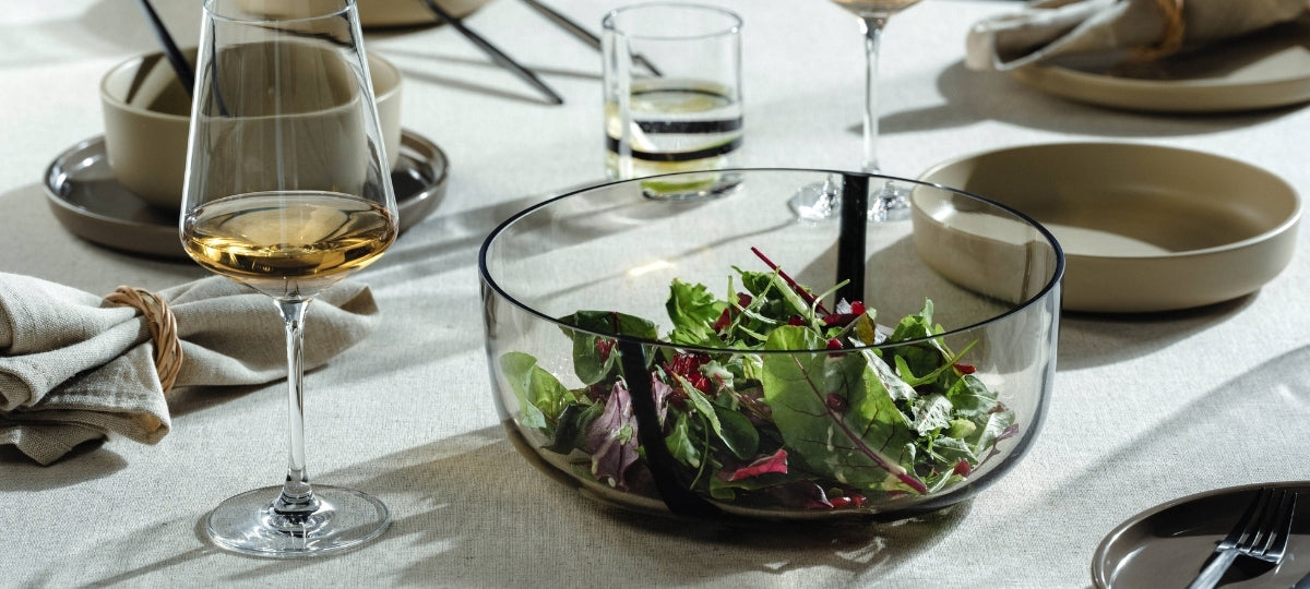 Elegantly set table with glass bowl of fresh salad, wine glasses with white wine, ceramic plates and linen napkins in neutral tones