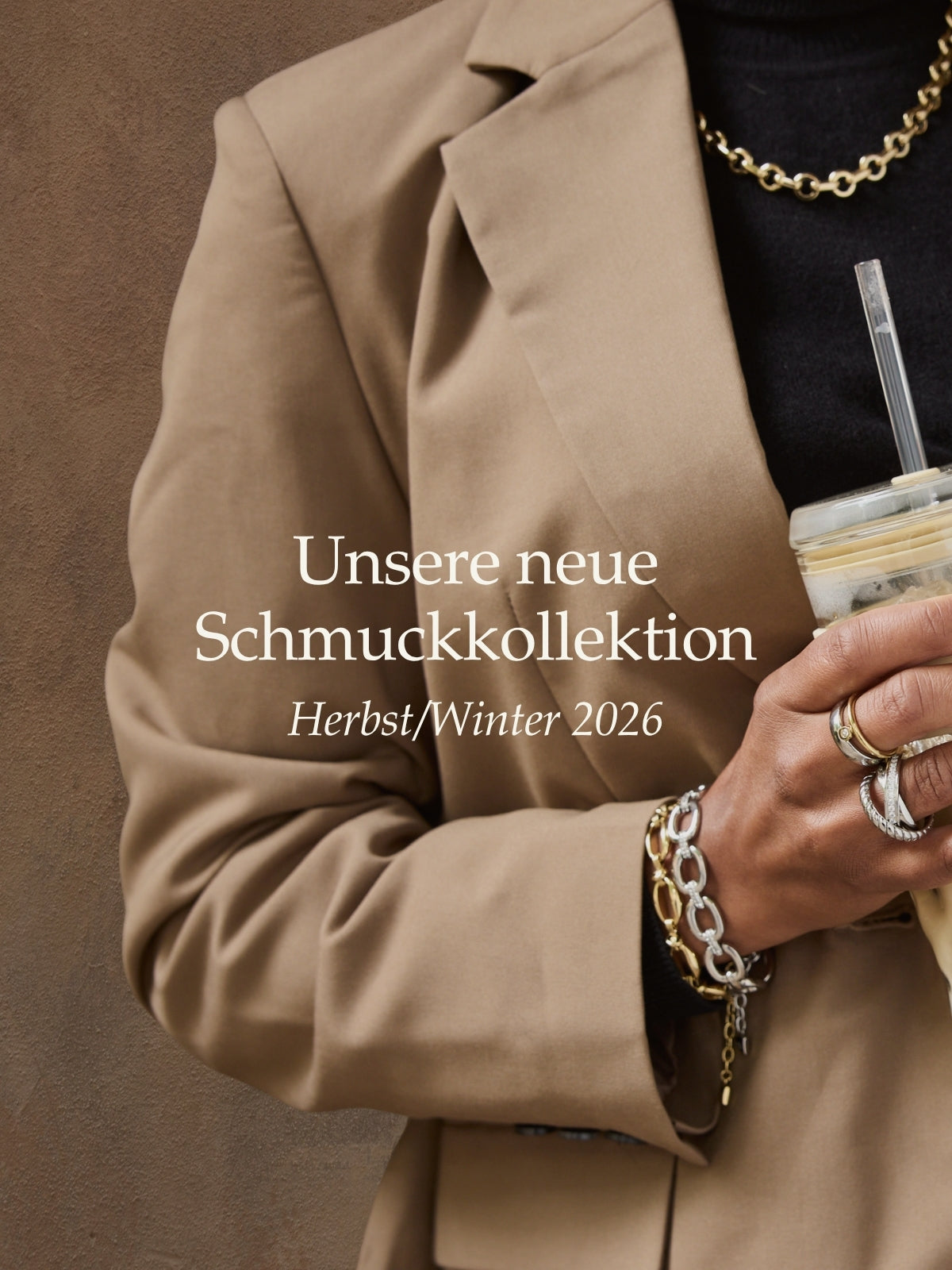 Unsere neue Schmuckkollektion Herbst/Winter 2026. Frau trägt beige Blazer, goldene und silberne Ketten, Ringe und hält Getränk mit Strohhalm