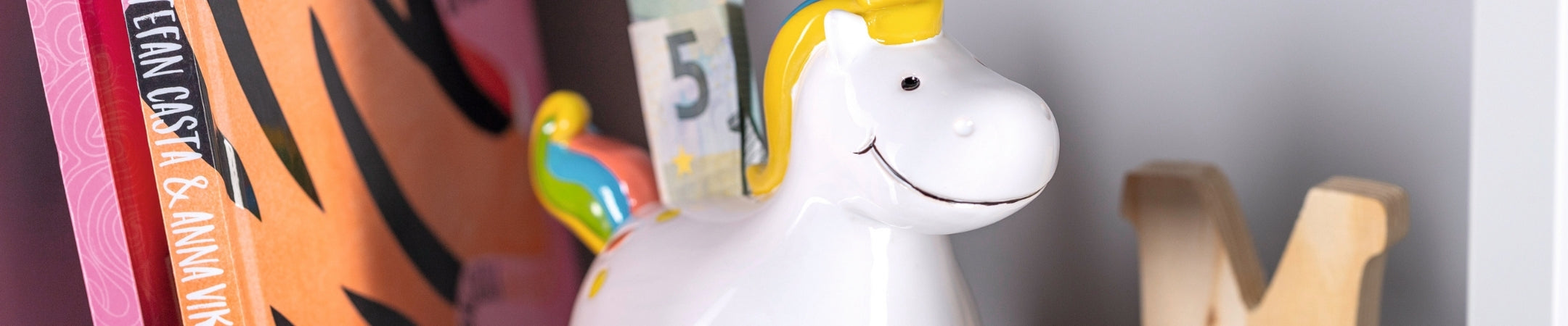 Weiße Spardose in Einhornform mit gelbem Horn neben bunten Büchern und Dekoobjekten