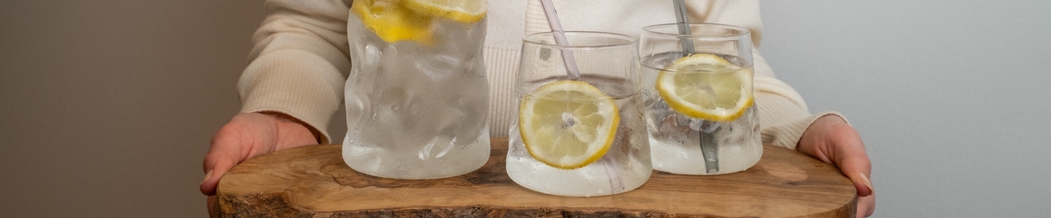 Hände halten Holzbrett mit vier Gläsern Zitronenwasser auf neutralem Hintergrund